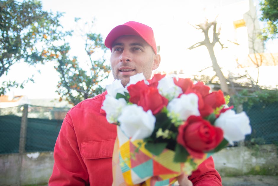 A man dressed in a red jacket and matching red cap holding a colorful bouquet of fresh flowers with red and white blooms, wrapped in decorative paper, outdoors in natural sunlight. The background feat