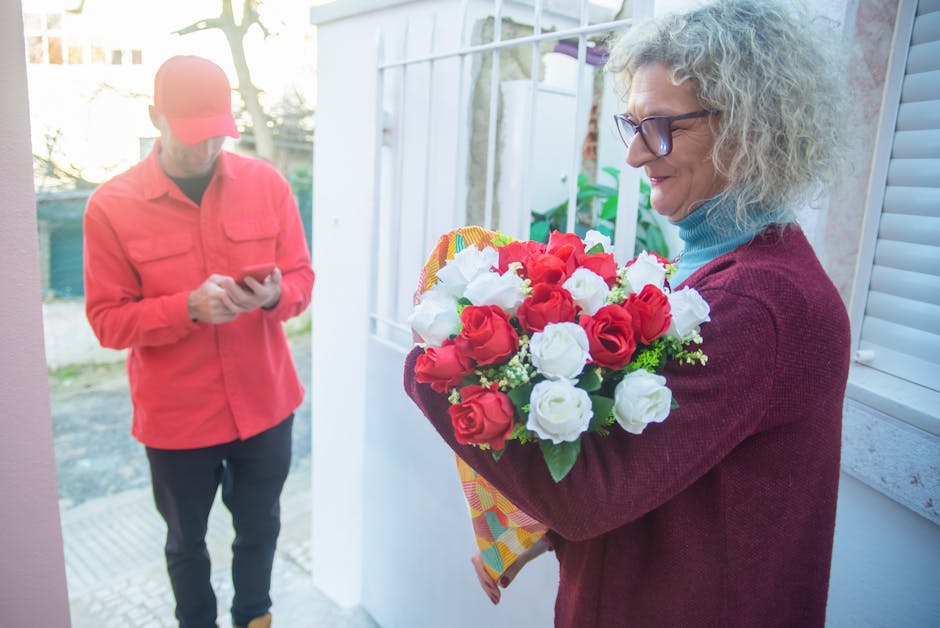 A woman with curly gray hair and glasses holding a vibrant bouquet of red and white roses wrapped in colorful paper, smiling as she receives a flower delivery. The flowers appear fresh with full petal