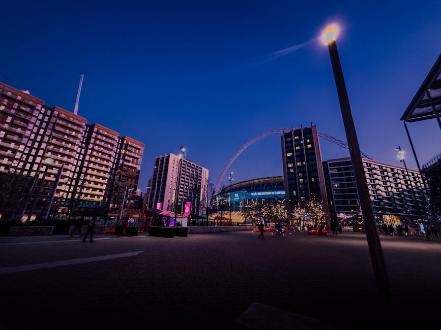 A nighttime cityscape featuring illuminated modern high-rise buildings around Wembley Park, with a brightly lit Ferris wheel in the background. The foreground shows a spacious pedestrian area with a f