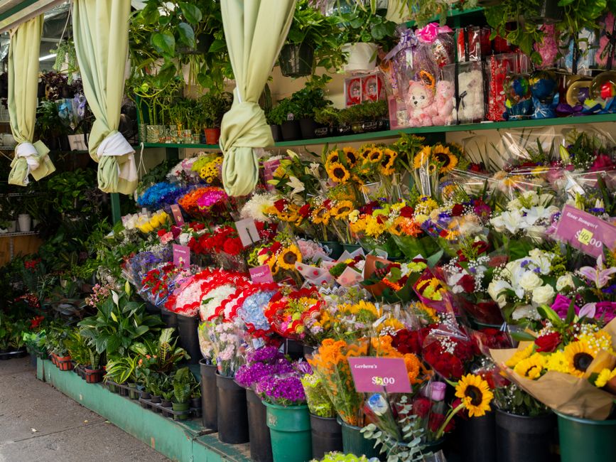 An outdoor flower shop display featuring a vibrant assortment of fresh flowers including sunflowers, roses, lilies, and carnations arranged in bouquets and wrapped in colorful paper and plastic. The f