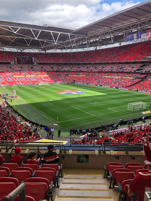 A view of Wembley Stadium's football pitch, featuring vibrant green grass with white marking lines, surrounded by red stadium seats filled with spectators. The seating area is covered by a modern roof
