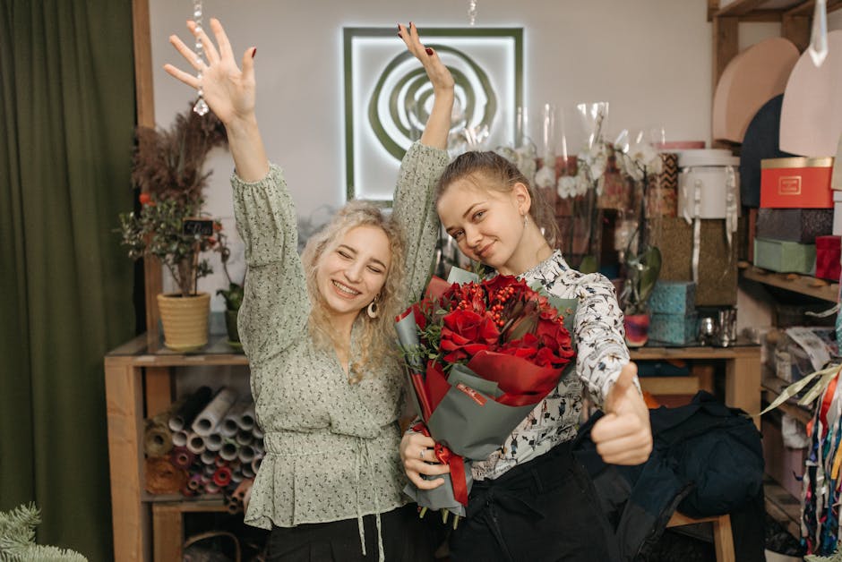 Two young women inside a flower shop, one holding a bouquet of vibrant red roses wrapped in grey and red paper, while the other celebrates with raised arms and a broad smile. They are surrounded by va
