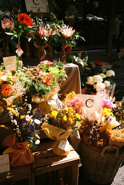 A display of fresh floral arrangements and bouquets on a wooden table and in wicker baskets, featuring a variety of flowers including pink lilies, red gerbera daisies, yellow chrysanthemums, white bab