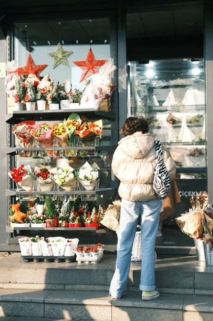 A woman stands outside a flower shop on Wembley High Road, looking at a display of vibrant floral arrangements. The shop window showcases a variety of fresh flowers, including bouquets wrapped in colo